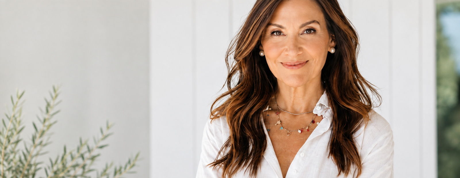 Smiling mature woman in a white blouse standing in a bright, naturally lit wellness-inspired setting with soft greenery in the background.